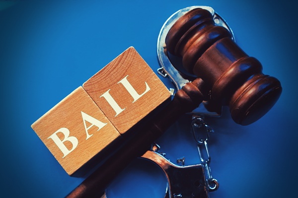 Wooden blocks spelling "BAIL" next to a judge's gavel and handcuffs on a blue surface.