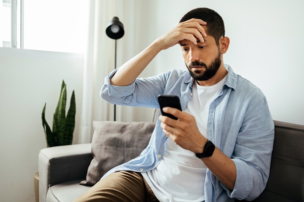 A man with a beard sitting on a couch, holding a smartphone and looking down with a stressed expression while resting his other hand on his forehead.