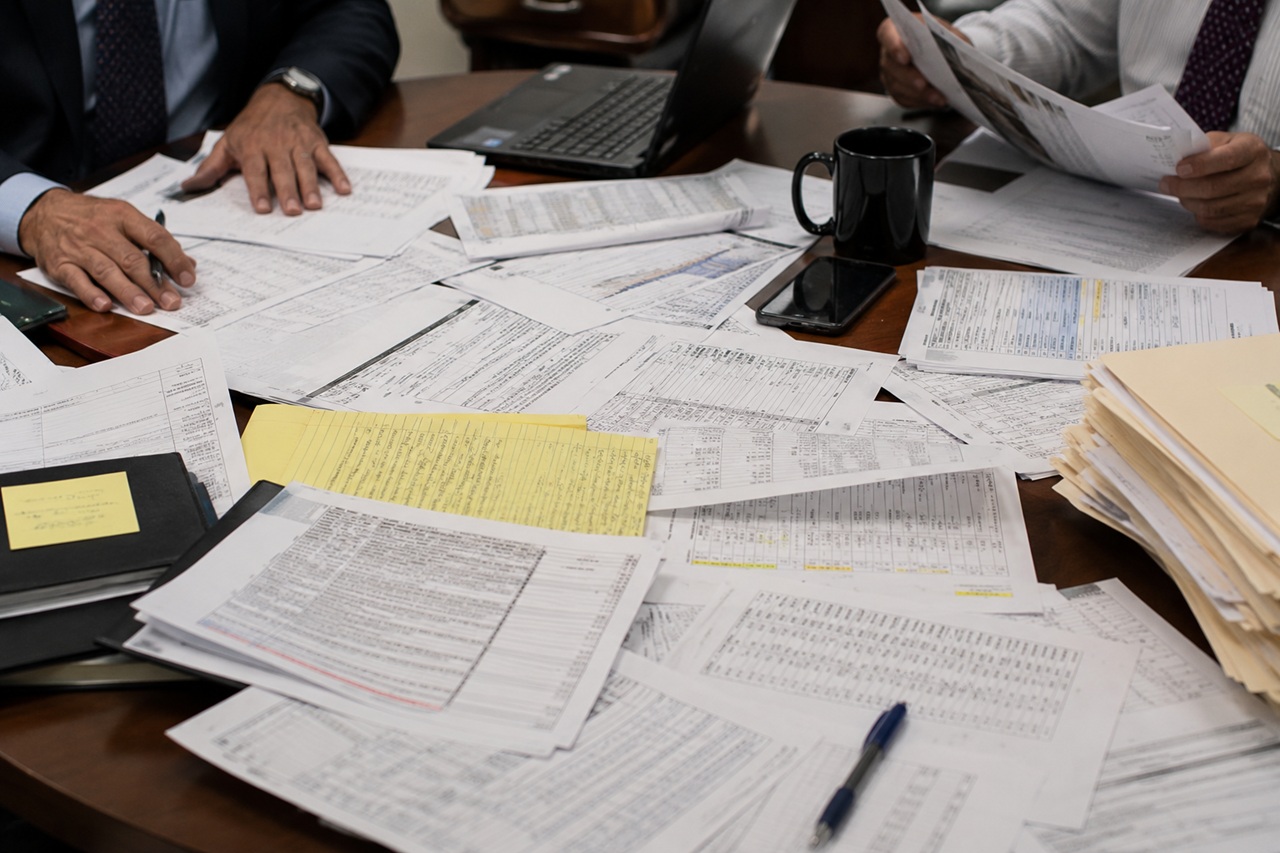 An overhead view of a wooden conference table cluttered with legal documents, financial spreadsheets, and investigation paperwork. Two professionals in business attire sit at the table; one reviews printed documents while the other has hands placed near sorted files. A black laptop, a smartphone, a black coffee mug, a blue pen, and a thick stack of manila folders are scattered across the table alongside yellow legal notepad sheets and sticky notes, suggesting a complex fraud investigation.